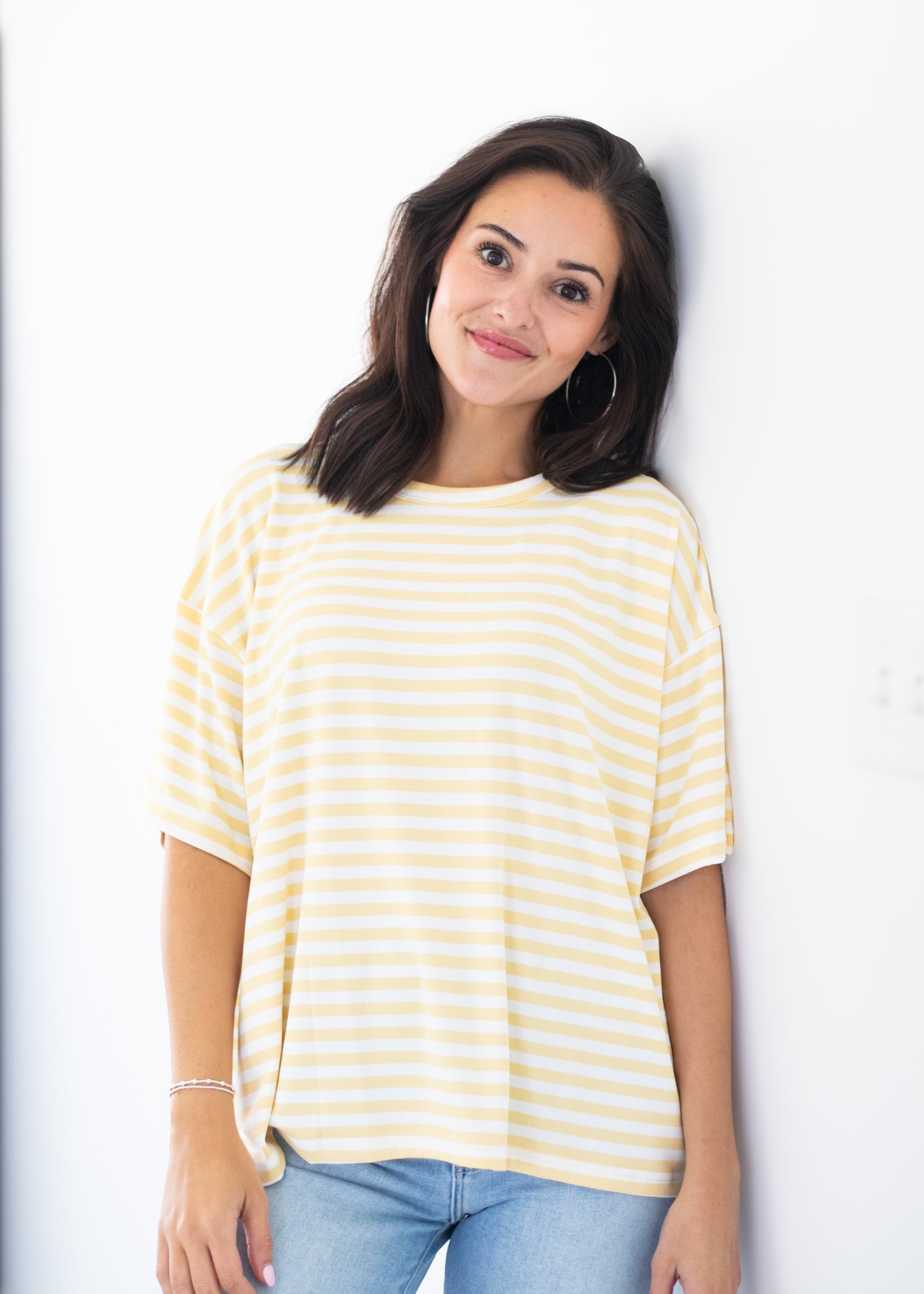 A woman with dark hair leans against a white wall wearing a pale yellow and white horizontally striped short-sleeve t-shirt and light blue jeans.