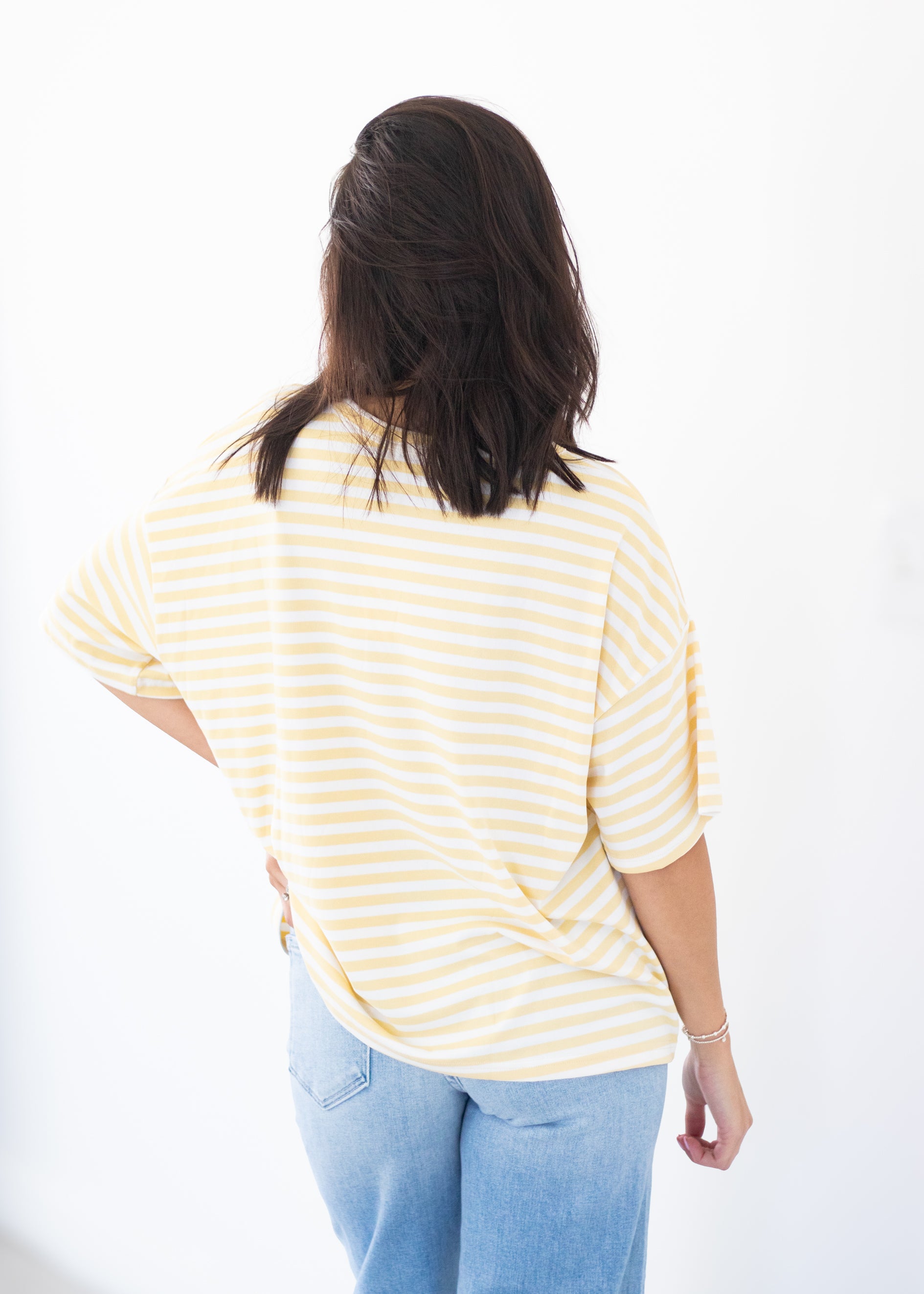A woman viewed from behind wearing a pale yellow and white striped short-sleeve top with light blue jeans, set against a plain white background.