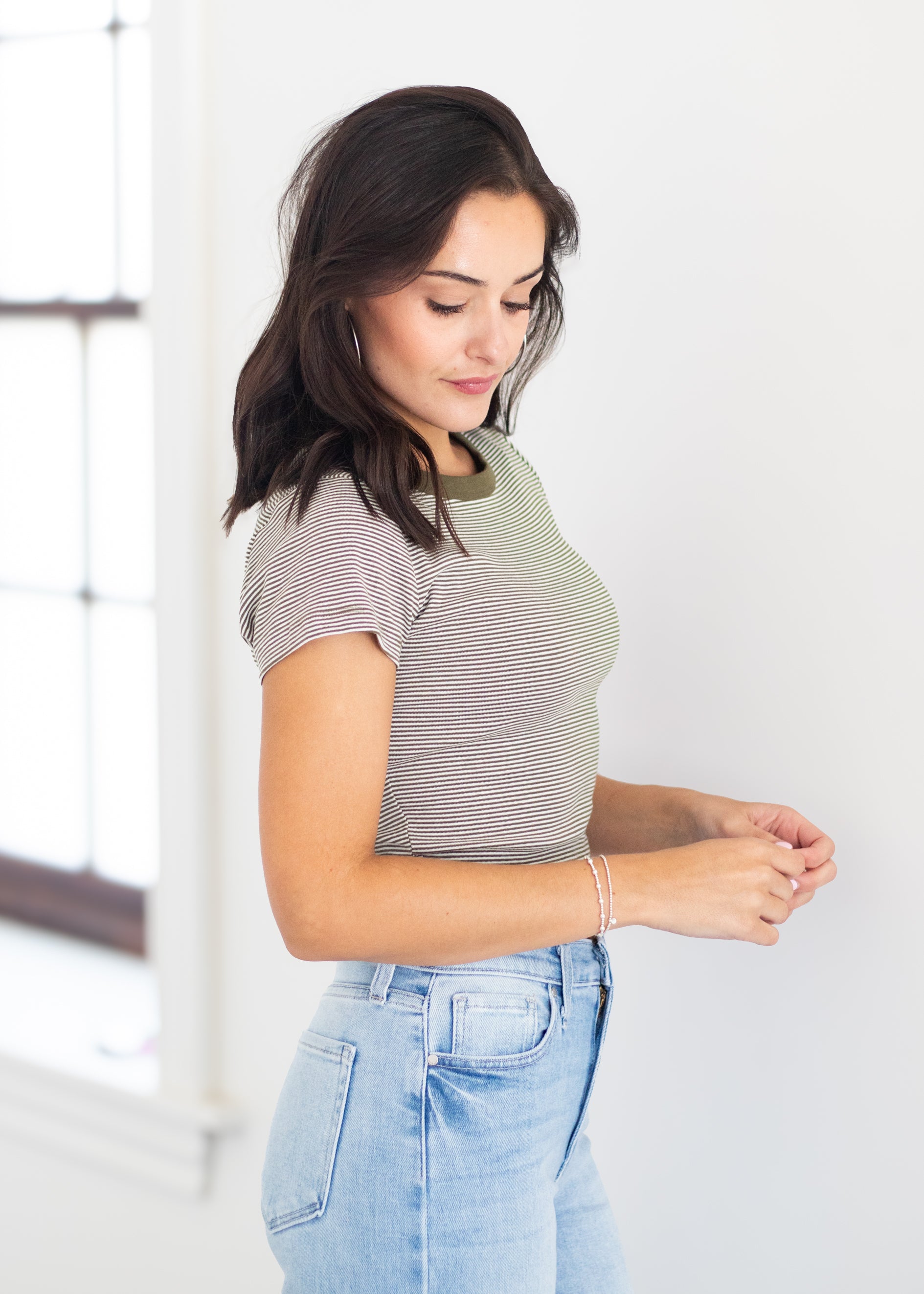 A woman wearing a short-sleeve crewneck t-shirt with thin gray and white horizontal stripes, paired with light blue jeans, standing in a brightly lit room.