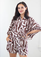 A woman modeling a matching brown and white zebra-print two-piece set consisting of a cropped button-up shirt and shorts, set against a plain white background.