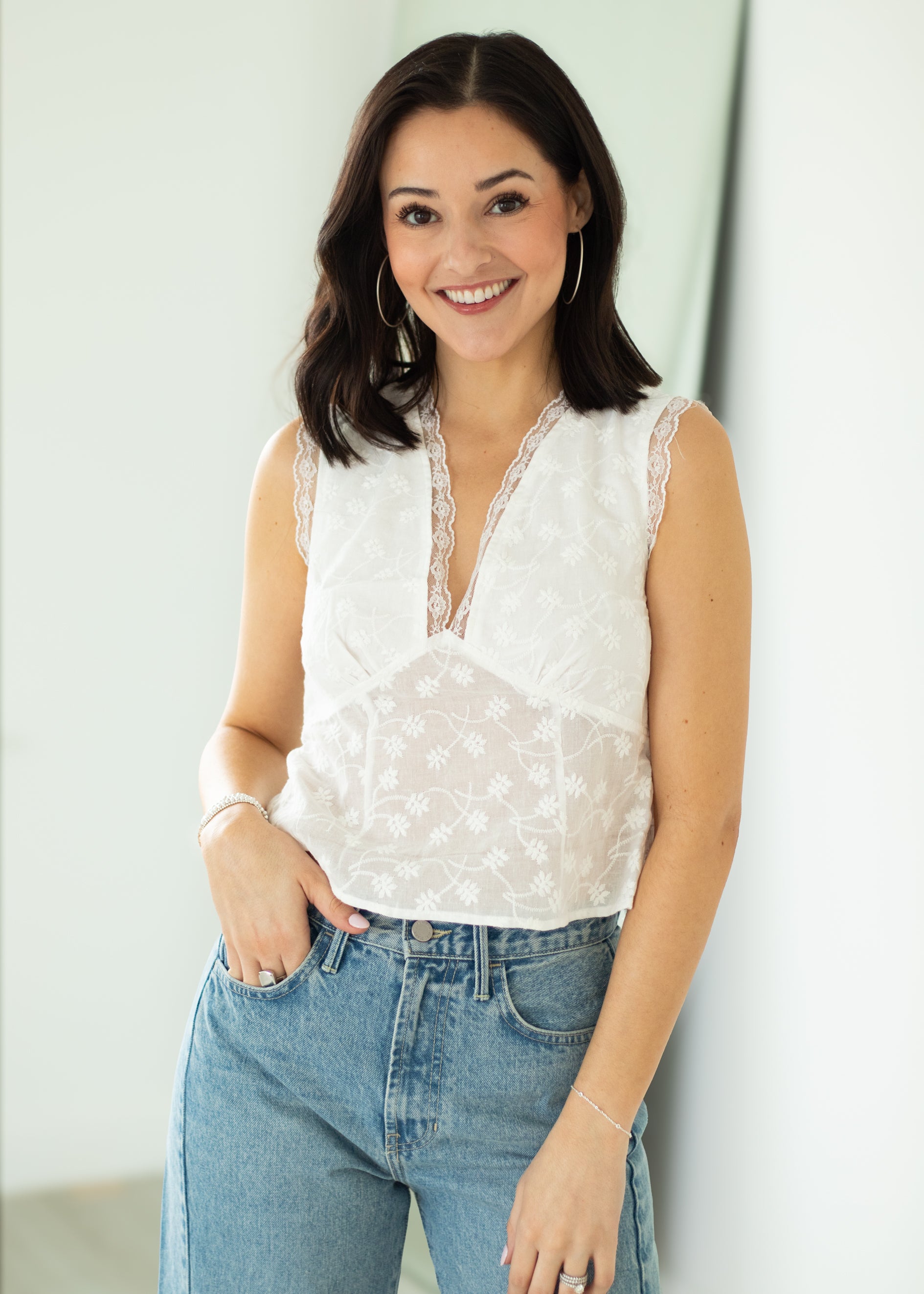 Woman wearing a white sleeveless top with lace details and blue jeans against a light background