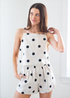 A woman modeling a two-piece linen romper set in cream with black polka dots, featuring a camisole top and matching shorts, set against a bright, neutral indoor background.