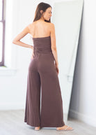 A woman wearing a strapless wide-leg jumpsuit in a muted brownish-purple shade, standing in a bright indoor room with a mirror in the background.