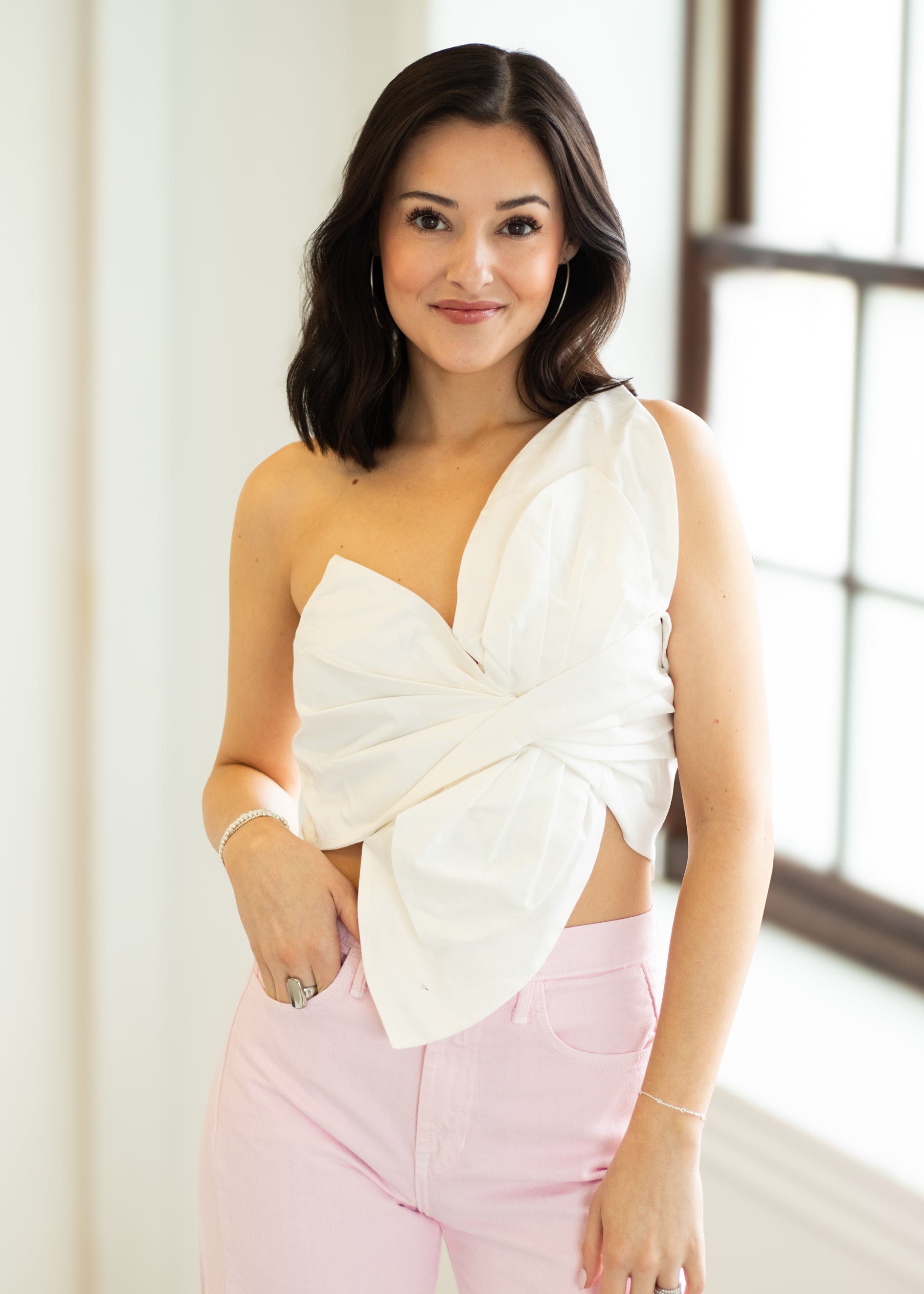 Woman wearing a white top with a bow detail and pink pants indoors.