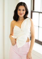 Woman wearing a white top with a bow detail and pink pants indoors.