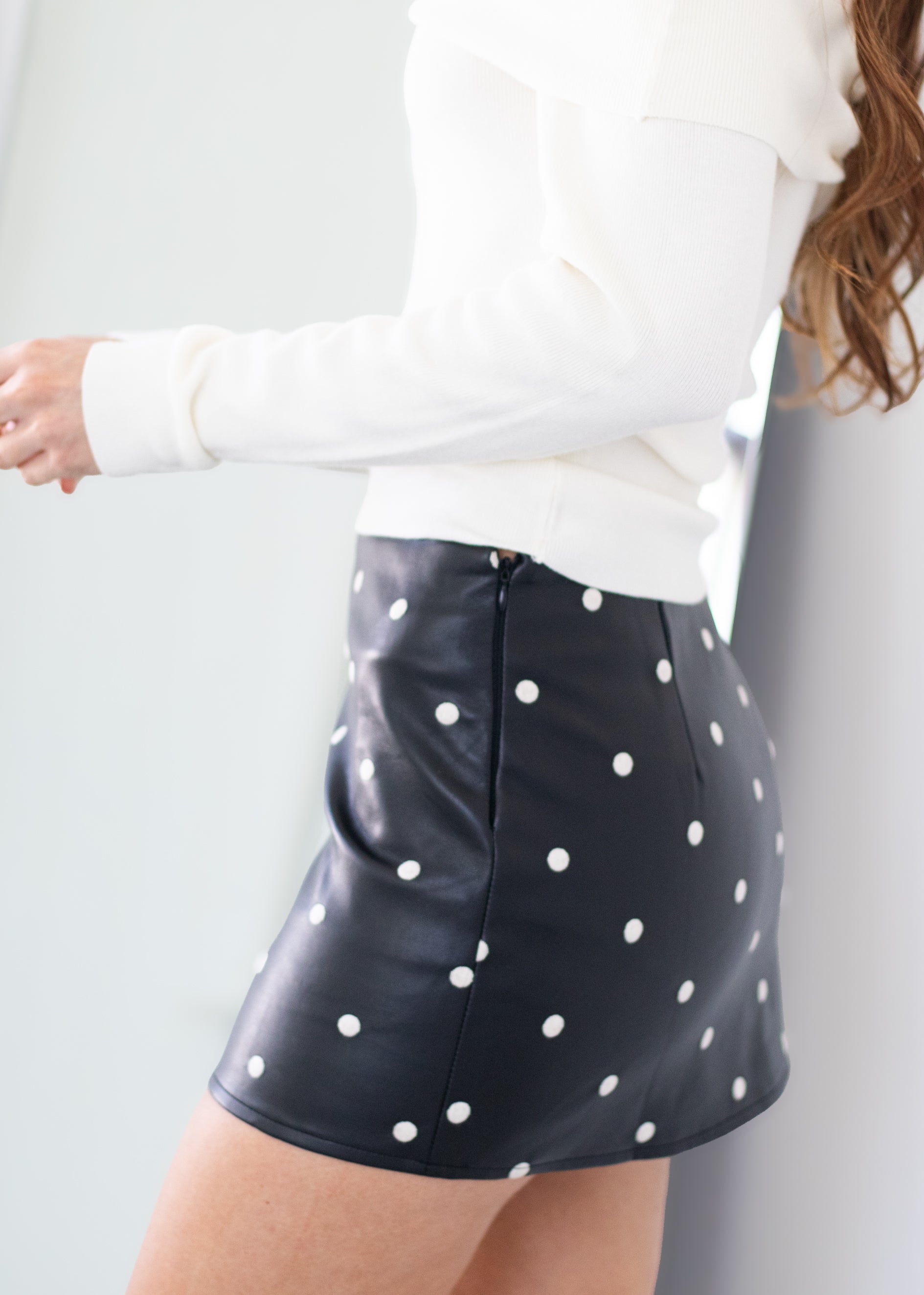 A model wearing a black faux-leather mini skirt with white polka dots, paired with a white long-sleeve top, shown from a side-back angle against a light gray background.