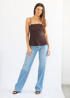 A woman modeling a dark brown camisole top with scalloped edges and light-wash blue jeans against a plain white studio background.