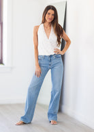A woman modeling a white sleeveless halter wrap top and light blue wide-leg jeans, standing in a bright, modern room with white walls and light wood flooring.