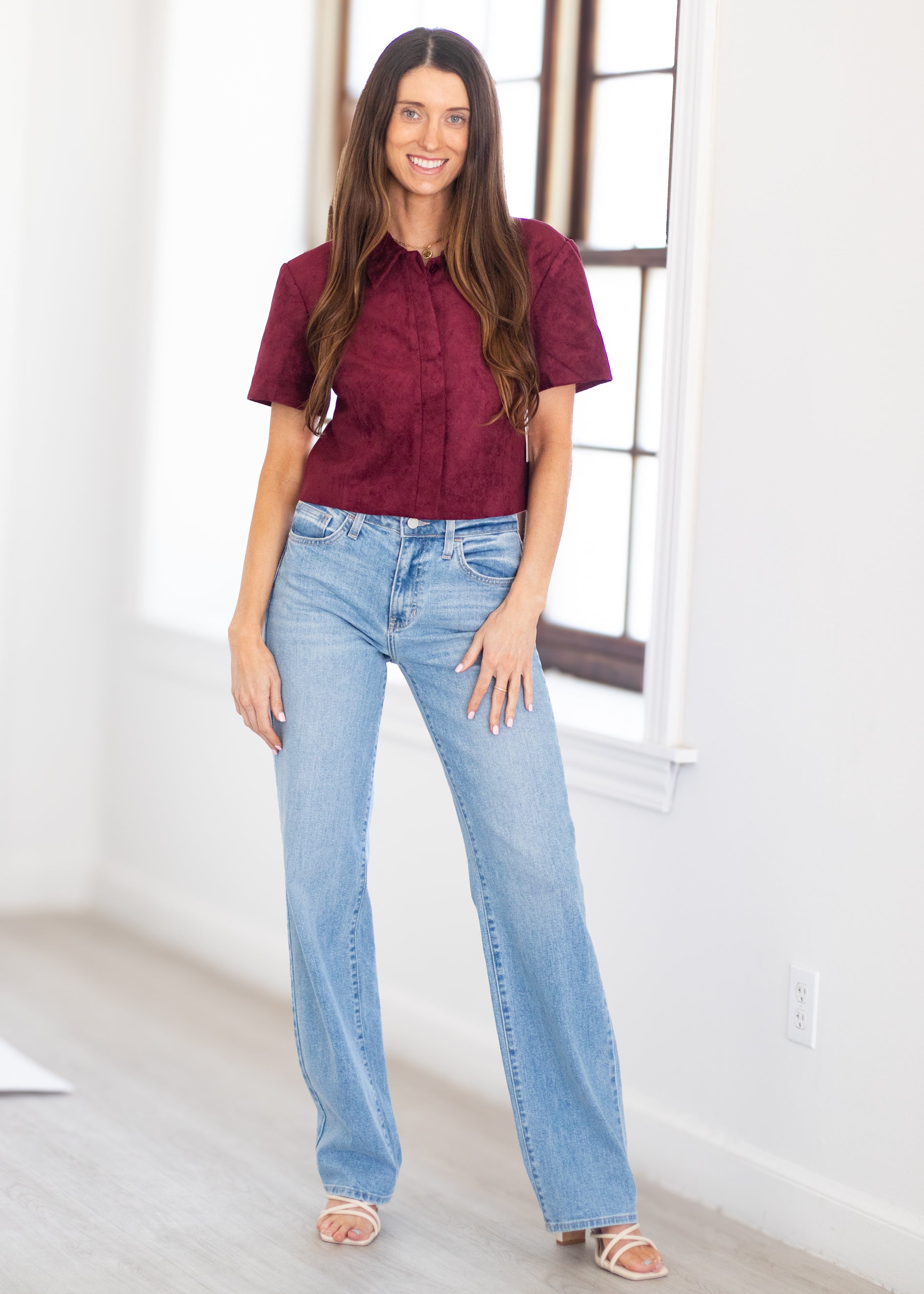 A woman modeling a short-sleeve burgundy button-up shirt and light-wash blue jeans, standing in a bright room with white walls and a window.