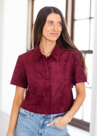 A woman wearing a short-sleeve collared button-up shirt in a textured burgundy or deep wine color, paired with light-wash blue jeans, standing in a bright indoor setting.