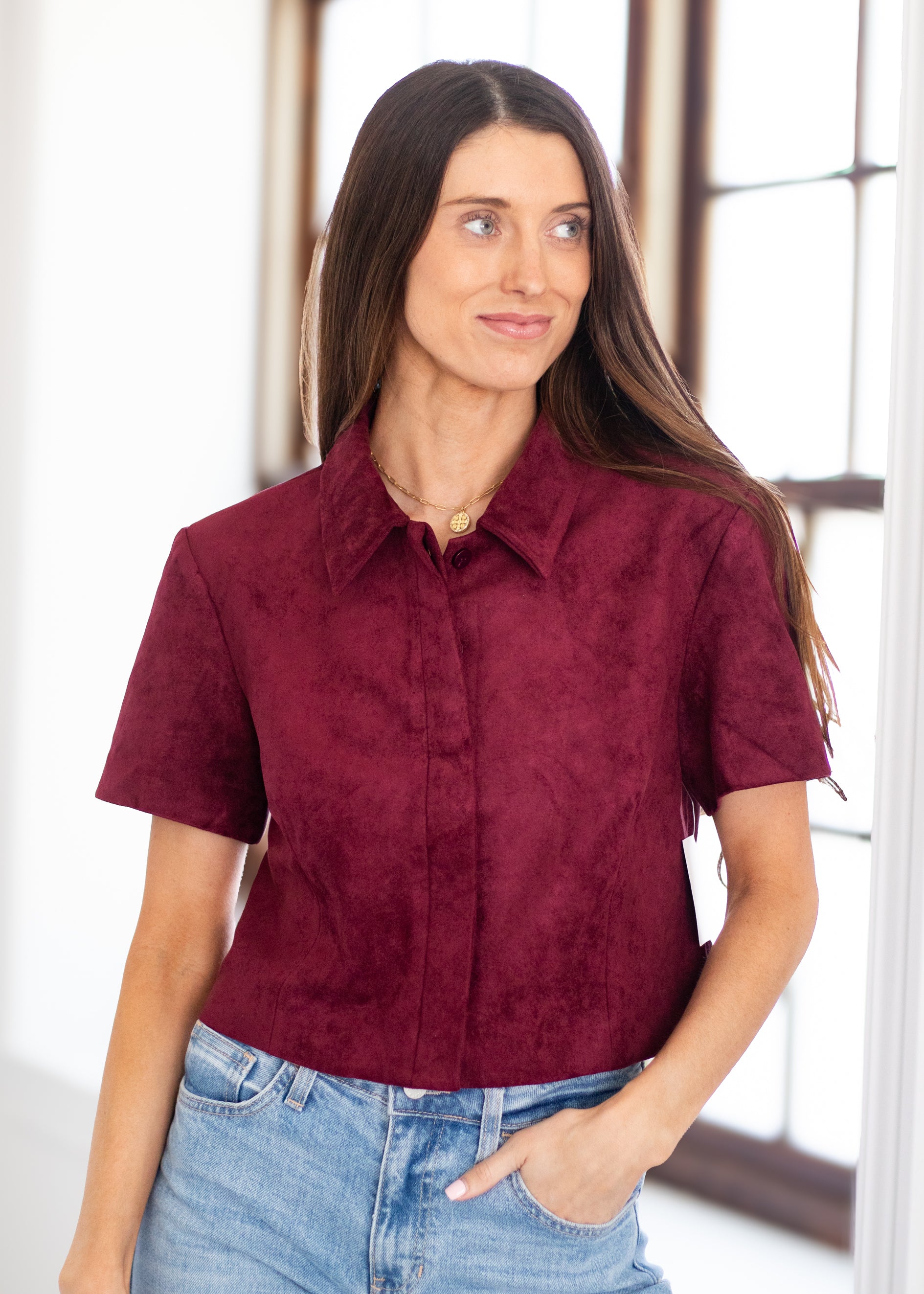 A woman wearing a short-sleeve collared button-up shirt in a textured burgundy or deep wine color, paired with light-wash blue jeans, standing in a bright indoor setting.