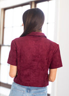A woman viewed from behind wearing a short-sleeve cropped jacket in a deep burgundy suede-like material, paired with light blue jeans, standing in front of a bright window.