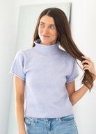 A woman with long brown hair wearing a light lavender short-sleeve mock neck knit sweater and blue jeans, standing in a brightly lit indoor setting.