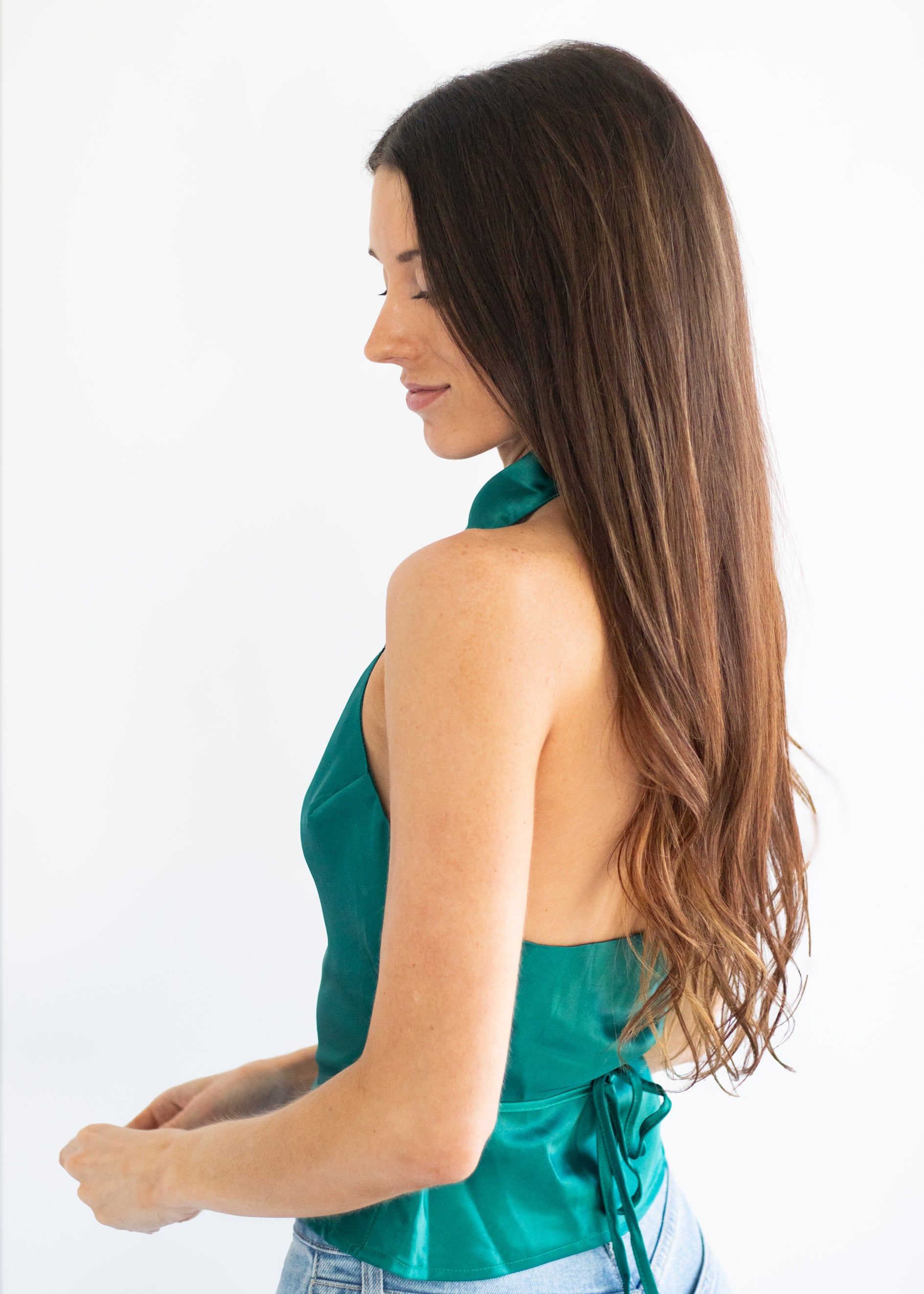 A woman with long brown hair is shown from the back wearing a satin-like teal green halter top and light blue jeans against a plain white background.