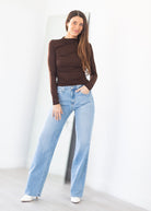 A woman modeling a brown long-sleeve ruched top and light-wash wide-leg jeans, standing in a bright white room.