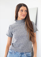 A woman with long brown hair wearing a gray short-sleeve mock neck knit sweater and blue jeans, standing against a light gray wall.