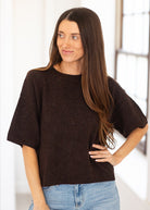 A woman with long brown hair wearing a dark brown short-sleeve knit sweater and light blue jeans, standing in a brightly lit indoor setting.