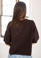 A woman viewed from behind wearing a dark brown short-sleeve knit sweater and light blue jeans, standing in a bright indoor setting near a window.