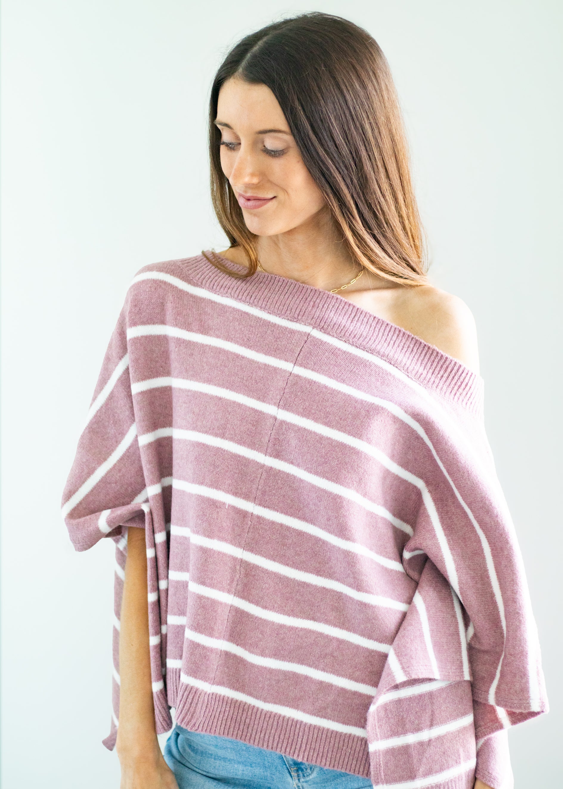 A woman wearing a dusty rose pink and white horizontally striped off-the-shoulder knit sweater, paired with blue jeans, against a plain light gray background.