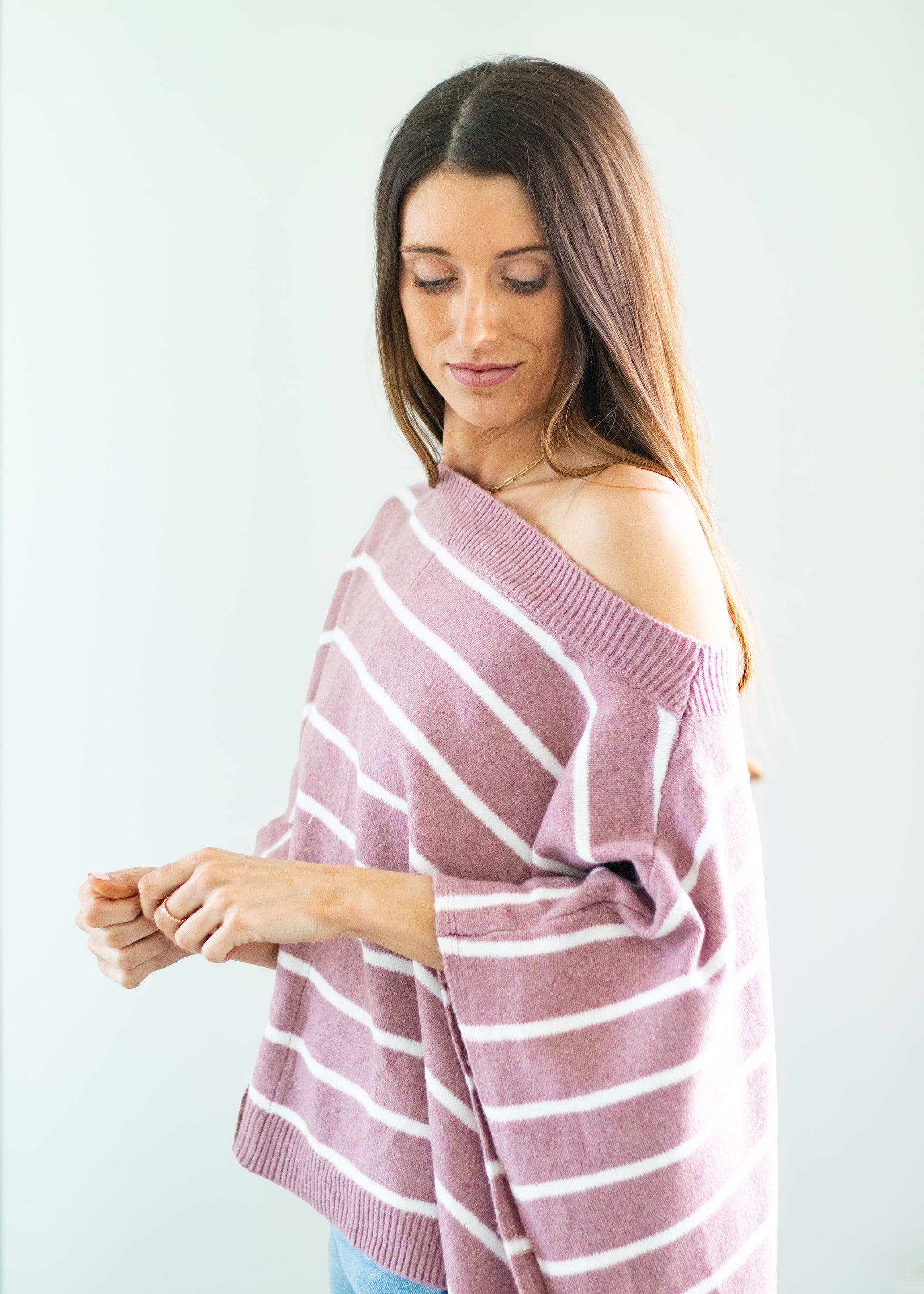 A woman wearing a dusty pink and white striped knit poncho sweater, shown from the side against a plain light gray background.