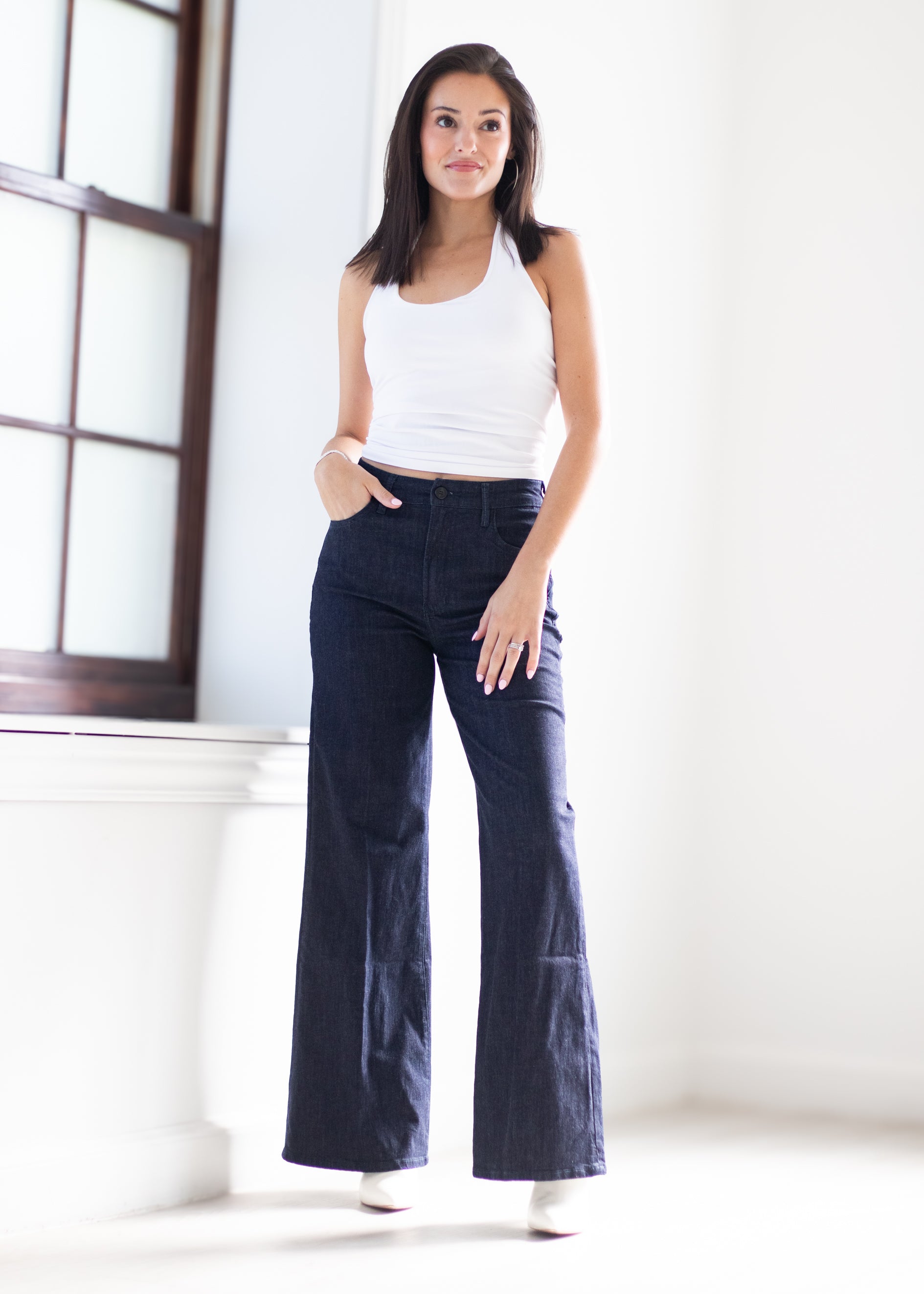A woman modeling dark blue wide-leg denim jeans with a white ribbed tank top, standing in a bright, minimalist room with a window on the left.