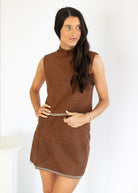 A woman modeling a sleeveless brown faux-suede mini dress with a mock neck and decorative checkered trim at the waist and hem, set against a plain white background.