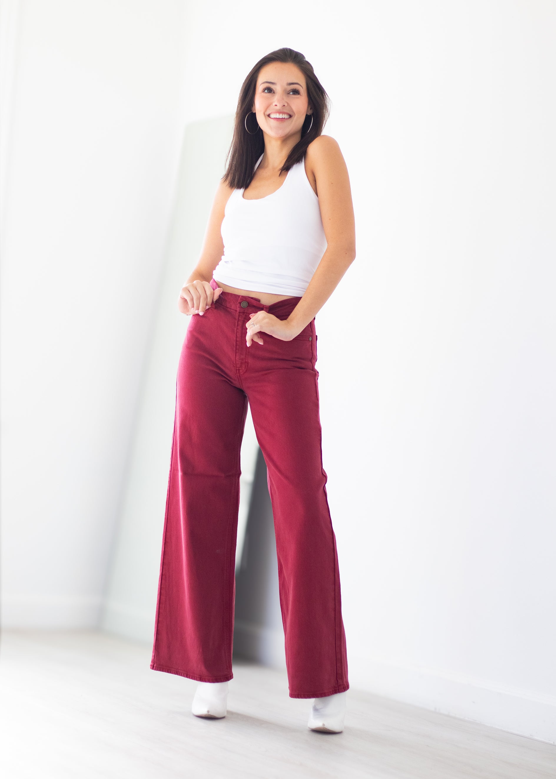 A woman modeling a pair of high-waisted, wide-leg burgundy pants with a white ribbed tank top, standing in a bright white room.
