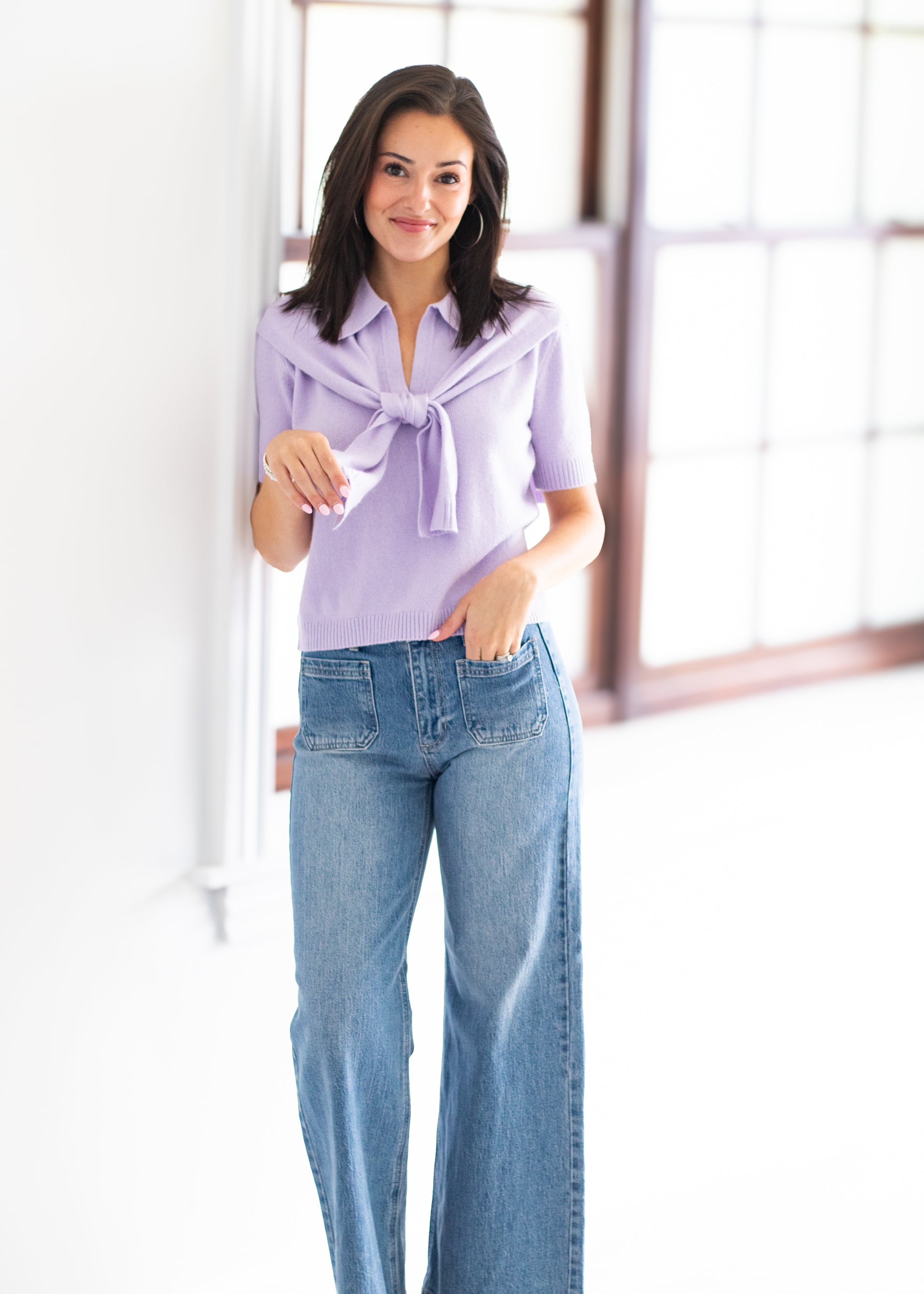 A woman modeling a light purple short-sleeve blouse with a tie-neck detail, paired with light-wash blue jeans, standing in a bright, modern room with large windows.