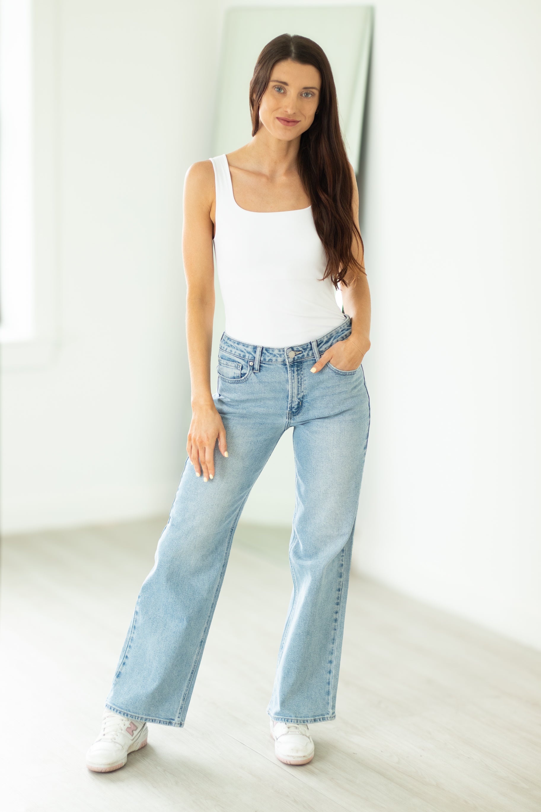 Woman wearing a white tank top and light blue jeans standing against a plain white background
