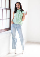 A woman stands in a bright white room wearing a mint green sleeveless blouse with a large bow at the collar, light-wash blue jeans, and white ankle boots.
