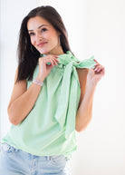 A woman wearing a light green and white vertically striped sleeveless top with a tie-neck detail, paired with light blue jeans, posing against a bright white background.