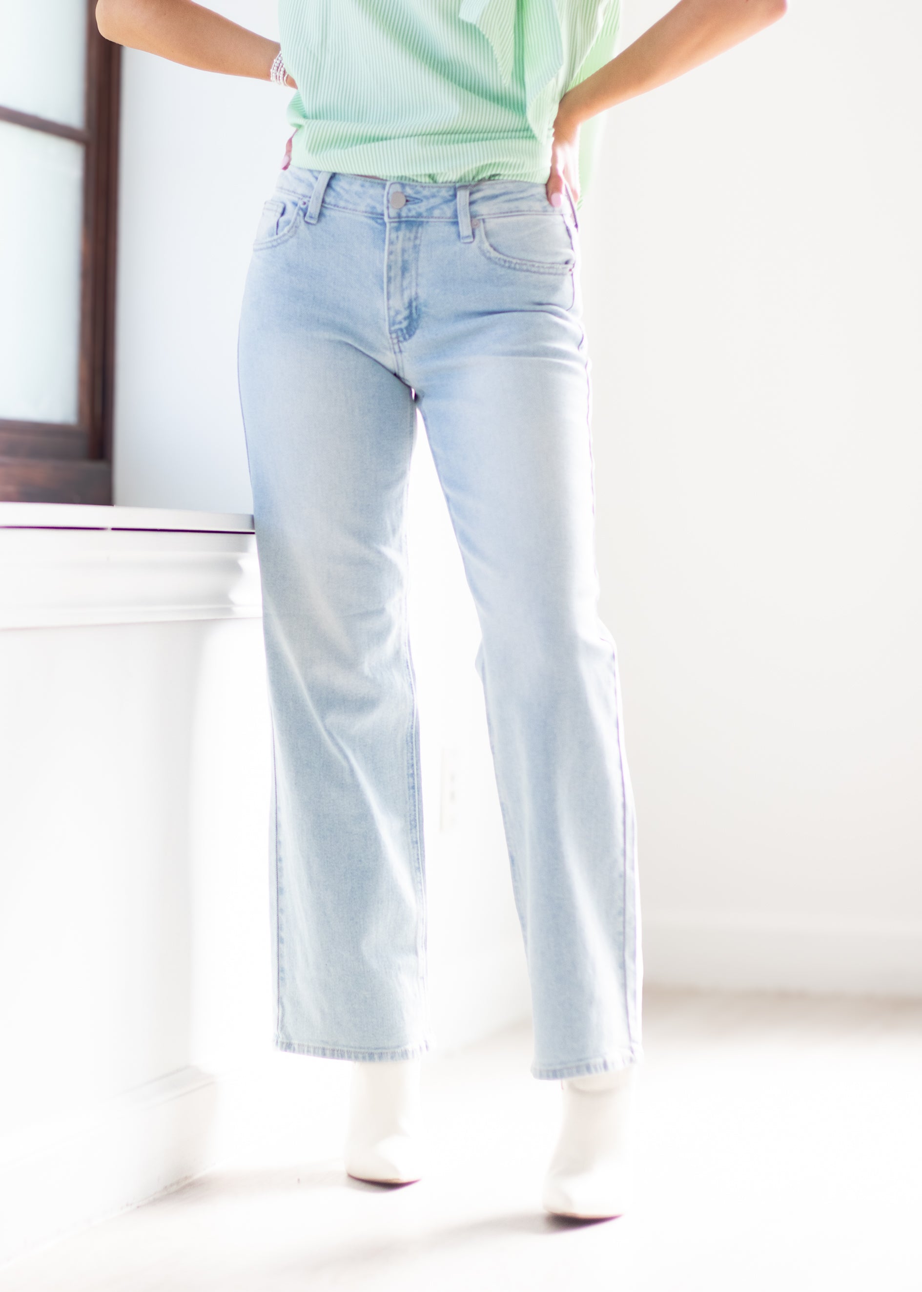 A person wearing light-wash blue straight-leg jeans, a mint green top, and white pointed-toe heels, standing in a bright white room.