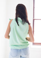 A woman viewed from behind wearing a sleeveless, ruffled high-neck top in light mint green and white vertical stripes, paired with light-wash denim jeans, standing in a brightly lit room.