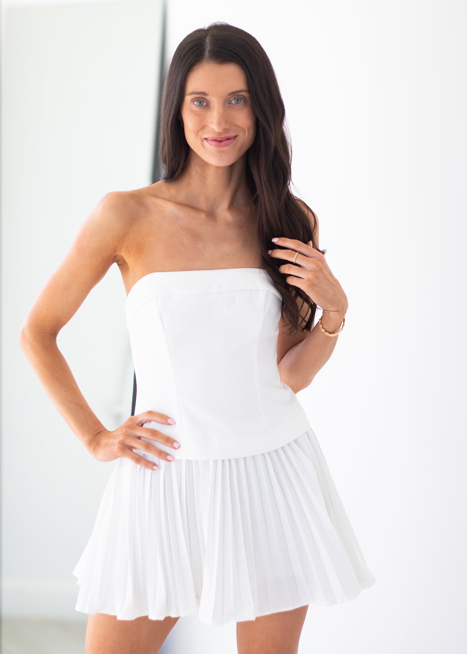 A woman modeling a white strapless mini dress with a structured corset-style bodice and a pleated flared skirt, posed against a bright white background.