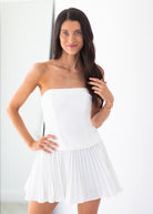 A woman modeling a white strapless mini dress with a structured corset-style bodice and a pleated flared skirt, posed against a bright white background.