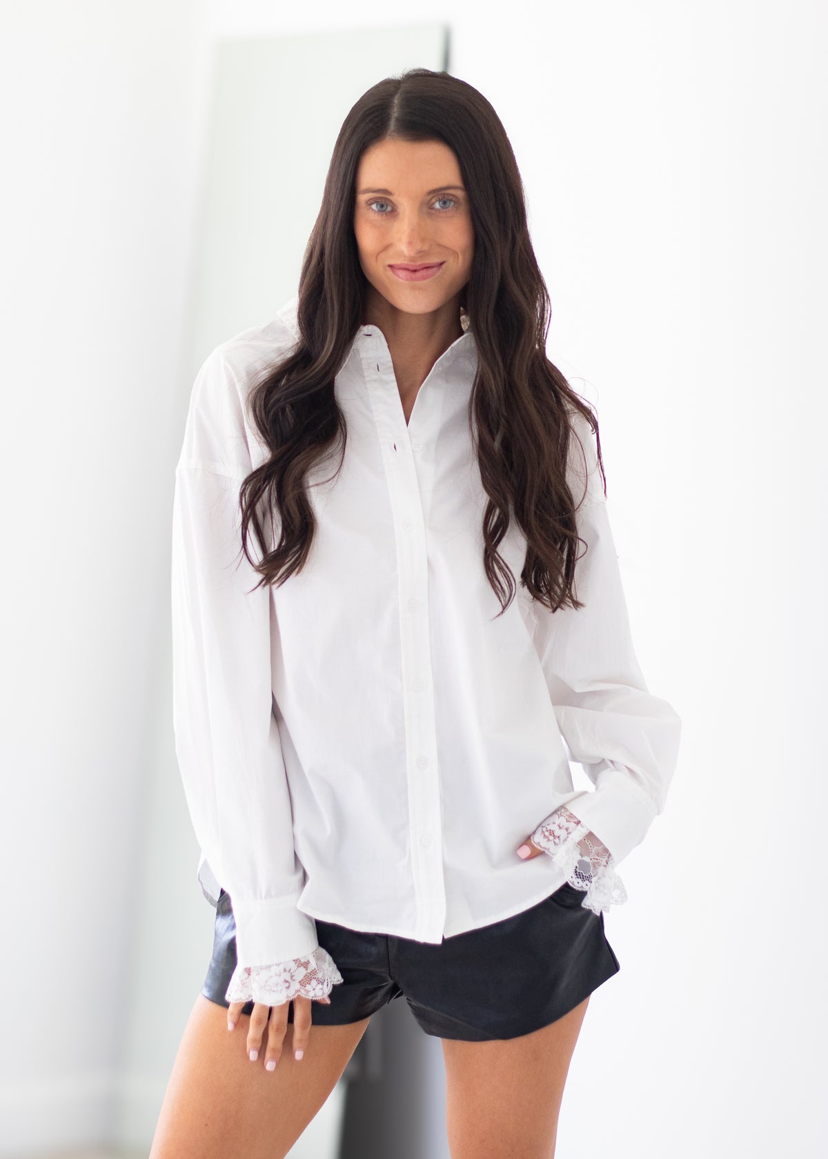 A woman with long dark hair wearing a white button-down shirt with lace cuffs and black shorts, standing against a bright white background.