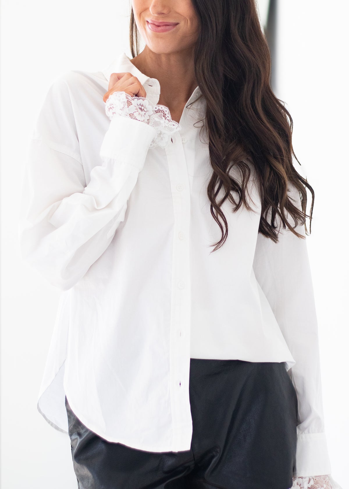 A woman wearing a white long-sleeve button-up blouse with delicate lace trim at the cuffs, paired with black pants, against a bright white background.