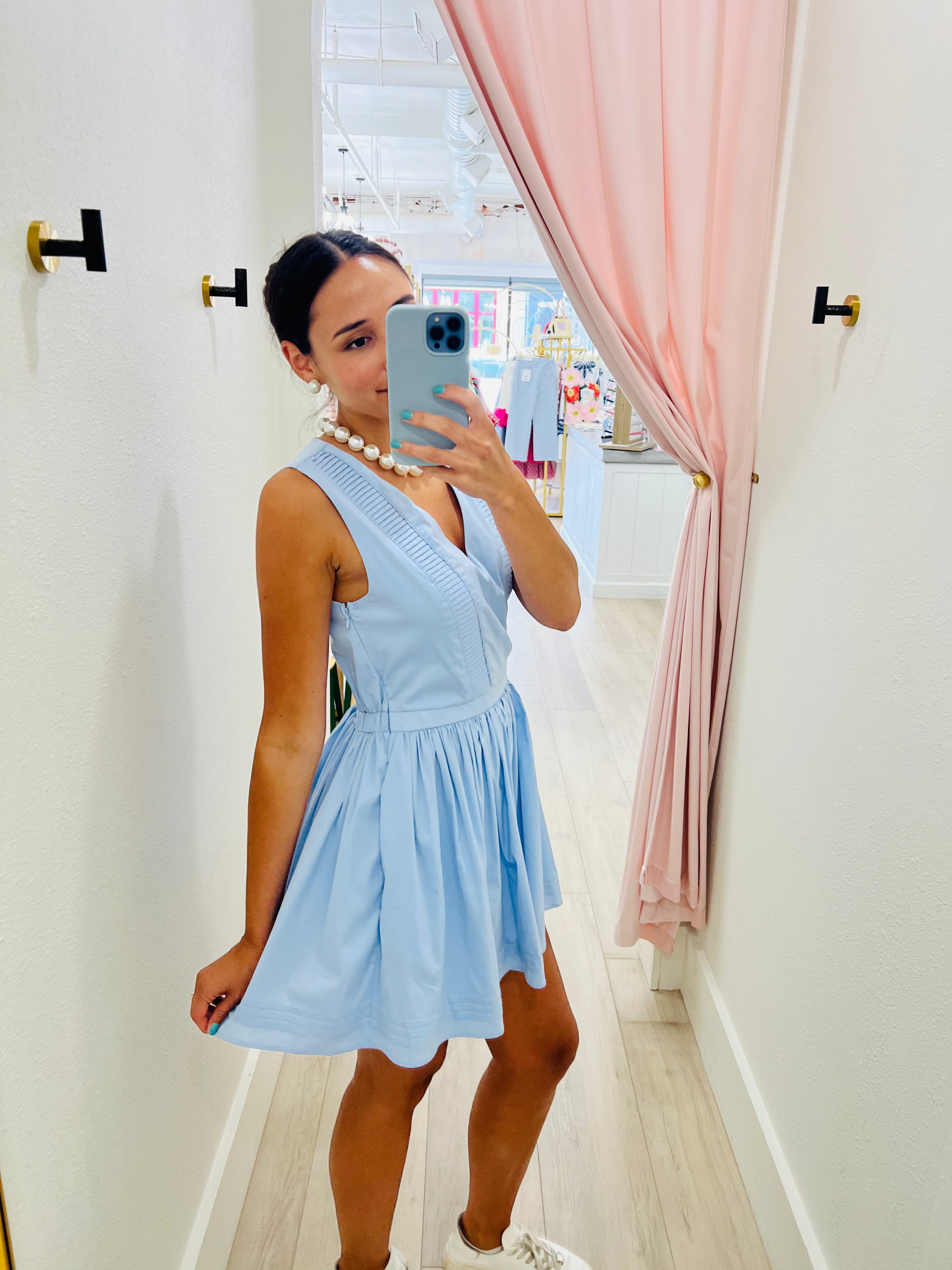 Woman taking a mirror selfie wearing a light blue dress in a room with pink curtains.