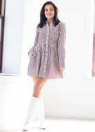 A woman models a long-sleeve mini dress with vertical stripes in brown and white, paired with white knee-high boots, standing in a bright, minimalist room.