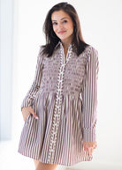 A woman modeling a long-sleeve collared dress with vertical brown and white stripes and a smocked bodice, photographed against a bright white background.