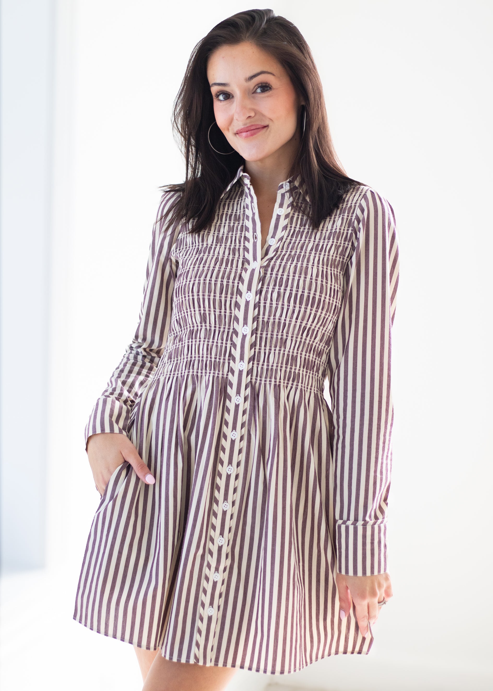 A woman modeling a long-sleeve collared dress with vertical brown and white stripes and a smocked bodice, photographed against a bright white background.