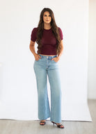 A woman modeling a short-sleeve burgundy knit top with a textured openwork pattern and light-wash blue wide-leg jeans, standing against a white studio backdrop.