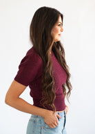 A woman with long, wavy brown hair is shown in profile wearing a short-sleeve knit top in deep burgundy and light blue jeans, set against a plain white background.