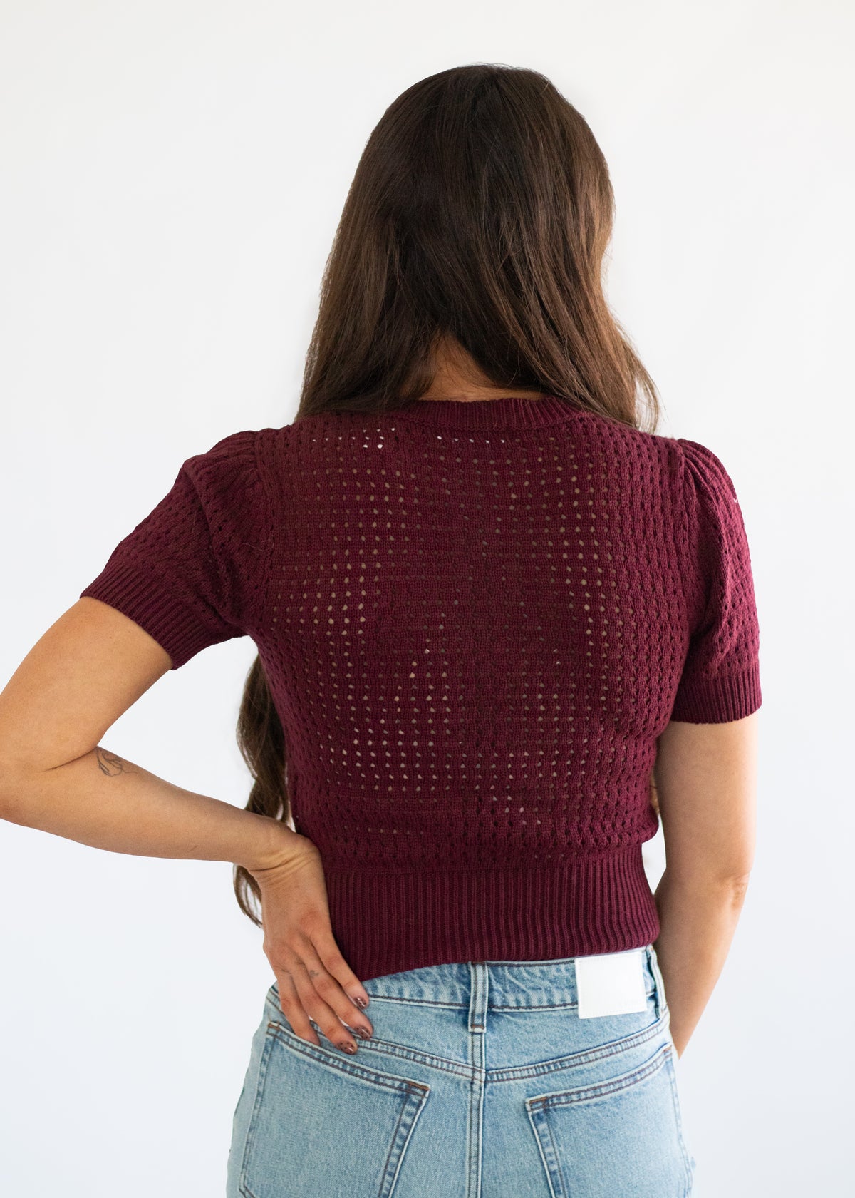 A woman viewed from behind wearing a short-sleeve maroon knit top with a textured openwork pattern and light-wash blue jeans against a plain white background.