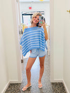 A woman takes a mirror selfie in a boutique wearing a light blue and white striped off-the-shoulder poncho top and matching striped shorts.
