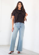 A woman modeling a dark brown knit wrap top and light-wash blue wide-leg jeans against a bright white studio background.