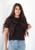 A woman with long brown hair wearing a dark brown short-sleeve knit sweater with a knotted front detail and light blue jeans, standing against a plain white background.