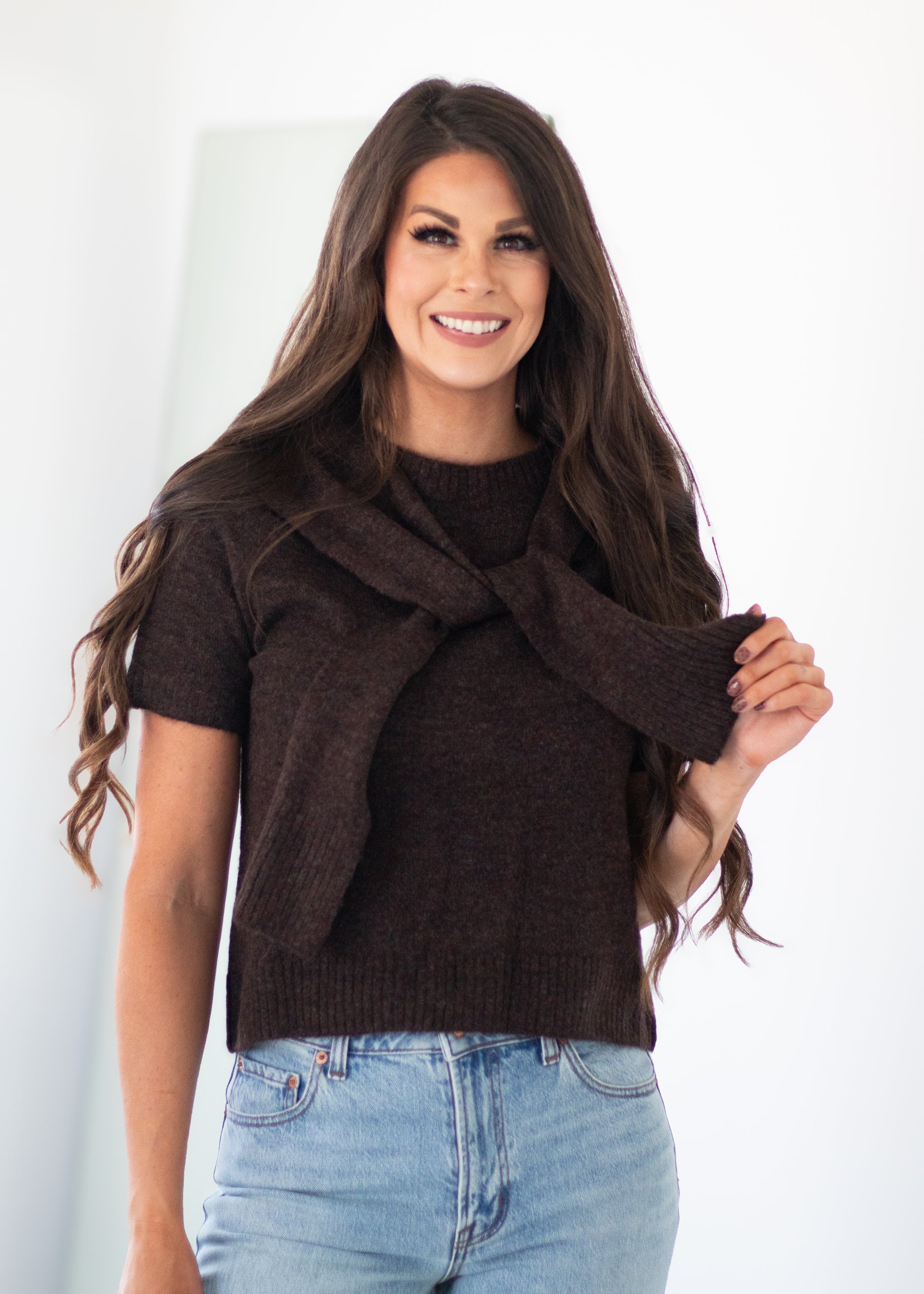 A woman with long brown hair wearing a dark brown short-sleeve knit sweater with a knotted front detail and light blue jeans, standing against a plain white background.