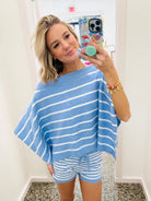 A woman takes a mirror selfie wearing a light blue and white horizontally striped knit poncho sweater and matching striped shorts.