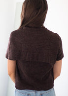 A woman viewed from behind wearing a dark brown short-sleeve knit top with a wide ribbed yoke and light blue jeans against a plain white background.